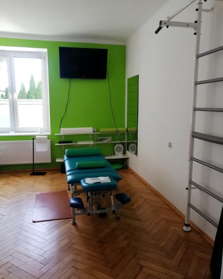 Sala zabiegowa gabinetu TULIKO: stół do masażu przy zielonej ścianie, drabinki gimnastyczne i okno